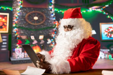 Santa Claus navigates web on his tablet gadget and checking gift lists and locations for delivery on world map. North Pole cabin filled with Christmas lights and magic, holiday preparations. clipart