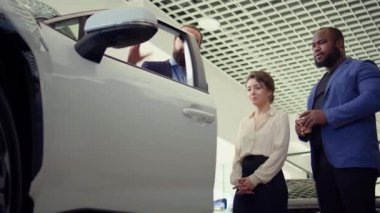 Araba galerisi satıcısı müşterilerden otomobil özelliklerini kontrol etmelerini ve mevcut modeller hakkında bilgi vermelerini istiyor. Ajan müşterilere araç kapısının dayanıklılığını gösteriyor, kamera B