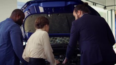 Satış elemanı kaputun altında müşteriler için parçalar gösteriyor, anlaşmaya varmayı umuyor. Showroom temsilcisi müşterileri satın almak için araç yapımında kullanılan parçaları, kamera B 'yi sunmaya ikna etmeye çalışıyor.
