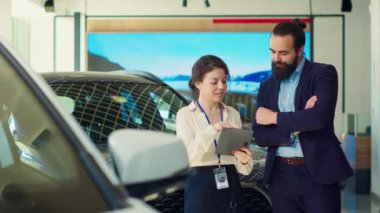 Satıcı kadın, tablet kullanarak adama seçenekler sunuyor. Satın alması için etki altına almaya çalışıyor. Showroom temsilcisi, aygıtta ayrıntıları gösteren, A kamerasında geniş güvence sunar.