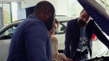 Araba kaputunun altındaki parçaları müşterilere gösteren dost canlısı araba satıcısı motor gücünü gösteriyor. Araç yapımında kullanılan parçaları sunan, müşterilerin otomobil seçmesine yardımcı olan ajan, kamera B