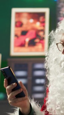 Dikey Noel Baba akıllı telefonuyla web 'de geziniyor ve dünya haritasında hediye listelerini ve teslimat yerlerini kontrol ediyor. Noel ışıkları ve büyüsüyle Kuzey Kutbu kabini, tatil hazırlıkları