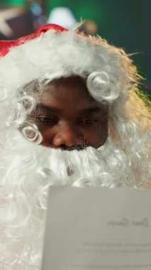 Dikey Video Baba Noel Baba, atölyesinde mektuplar ve listeler okuyor, beyaz sakallı adam posta kutusunu açıyor. Tatil ışıkları ve süslemelerle çevrili Noel Baba gelenek için hediyeler hazırlar.