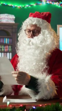 Neşeli Noel Baba 'nın dikey portresi ateşin yanında mektuplar okuyor, çelenklerle süslenmiş neşeli bir ruh ve kamarasında parıldayan ışıklar. Noel Baba, Kuzey Kutbu 'nda oyuncak dilekleri kabul ediyor.
