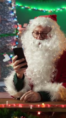 Dikey Noel Baba akıllı telefonuyla web 'de geziniyor ve dünya haritasında hediye listelerini ve teslimat yerlerini kontrol ediyor. Noel ışıkları ve büyüsüyle Kuzey Kutbu kabini, tatil hazırlıkları