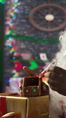 Dikey Video Baba Noel Baba ev yapımı zencefilli kek evini şekerleme ve boncuklarla süslüyor, Kuzey Kutbu kabin atölyesi. Siyah Noel Baba, tarçınlı tatlı pişirme geleneğini devam ettiriyor.