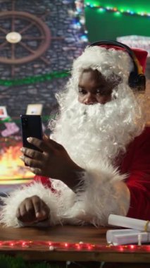 Dikey Videolu Neşeli Aziz Nicholas cep telefonundan Noel şarkıları dinliyor, takım elbiseli siyah Noel Baba kış tatili ruhunu kütük gibi kabinine yayıyor. Dijital teknoloji