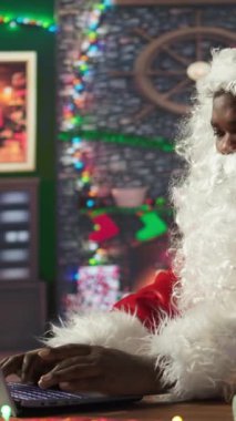 Dikey Video Siyah Noel Baba, Kuzey Kutbu 'ndaki şenlikli bir kulübede dizüstü bilgisayarını kullanarak hediye yeri arıyor. Aziz Nicholas dünyaya bağlı, Noel büyüsünü gerçekleştiriyor. Kamera B.