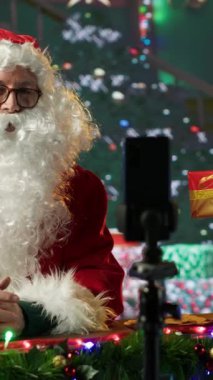 Kırmızı takım elbiseli Dikey Video Aziz Nicholas, süslü Kuzey Kutbu kulübesinde video günlüğü çekiyor. Beyaz sakallı nüfuzlu kişi bayram geleneklerini izleyicilerle paylaşıyor.