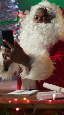 Dikey Video Siyah Noel Baba, Kuzey Kutbu atölyesindeki bir telefona cevap veriyor ve Noel arifesi için hazırlık yapıyor. Kırmızı elbiseli beyaz sakallı adam kış geleneğini vurguluyor.