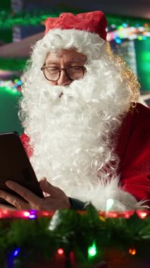 Dikey Video Baba Noel Baba, çevrimiçi hediye listesini rahat kütük kulübesindeki bir tabletten kontrol ediyor. Kuzey Kutbu 'ndaki adam internetten dünyaya bağlanıyor ve Noel büyüsünü hayata geçiriyor.