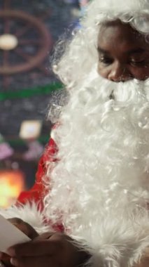 Dikey Video Siyah Noel Baba, Kuzey Kutbu 'ndaki rahat kulübesinde, x noel şenliği için süslü dekorasyonlarla çevrili Noel mektuplarını okuyor. Gelenek ve sihir bayram neşesini canlandırır