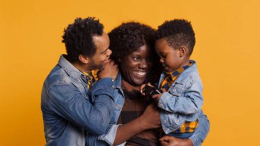 Küçük neşeli çocuk video oyunları için bir kontrolörle oynuyor, anne ve babasıyla stüdyoda poz veriyor. Sarı arka planda küçük çocukları olan Afro-Amerikan bir aile, çocuk oyunları. Kamera A.