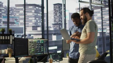 IT employees in office working together using laptop, testing and deploying programs and systems. Developer and colleague checking code on notebook, doing brainstorming in workspace, camera B clipart