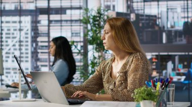 Kadın yönetici masasında iş operasyonlarını inceliyor, proje yönetimine odaklanmış. Çalışan, girişim stratejisini optimize etmek için performans ölçümlerini ve bütçe tahsislerini analiz eder. Kamera A.