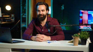 Happy etkili mikrofon kullanıyor, loş ışıklandırılmış stüdyo içine giriyor, hayranlarıyla sohbet ediyor. Apartmanın başlangıç akışında podcast kayıt ekipmanlarıyla çevrili gülümseyen adam, kamera A