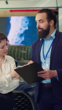 Kullanılabilir modeller, özellikler ve fiyatlandırma seçenekleri hakkında bilgi sağlayarak müşterilere panodaki metni okuyan dikey video araba bayii satıcısı. Temsilci müşterilerle araç fiyatlarını görüşüyor