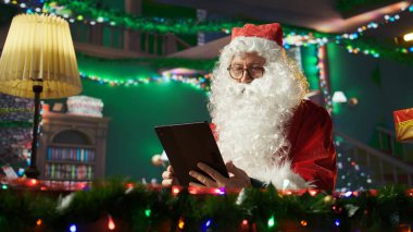 Father Christmas checking his online gift lists on a tablet in his cozy log cabin. Man at the North Pole connected to the world through the internet, bringing Christmas magic to life. Camera A. clipart