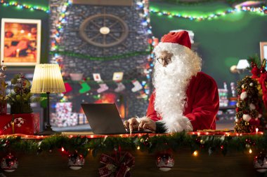 Father Christmas is browsing websites on his laptop and checking holiday gift lists from his North Pole cabin. The festive season is alive with online magic and holiday traditions. clipart