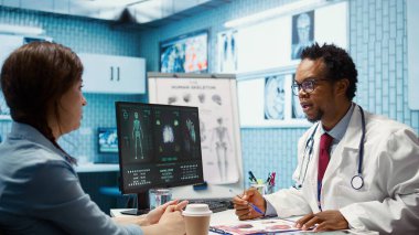 Ofiste röntgen görüntülemesi ve test bulgularını inceleyen profesyonel doktor kontroldeki hastaya tanı raporu veriyor. Afrika kökenli Amerikalı uzmanlar yorum için taramaları inceliyor. Kamera B.