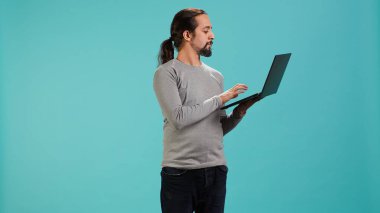 Bilgisayarda vakit geçiren gülümseyen kişinin portresi, stüdyo arka planında izole edilmiş. Neşeli adam not defterindeki sosyal medya paylaşımlarını kontrol ediyor, boş zamanların tadını çıkarıyor, kamera A