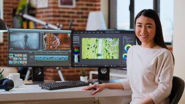 Asyalı kadın video editörü oturma odasında çift monitörlerle çalışıyor, video görüntülerini düzenliyor ve modern ofis ortamında renk sınıflandırması yapıyor. Prodüksiyon ve medya yaratımı için mükemmel. Kamera B.