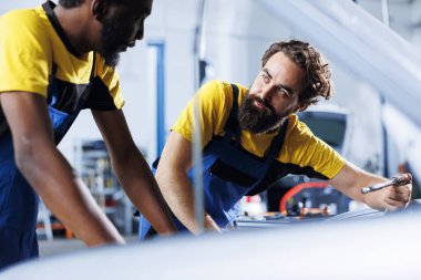 Technicians collaborating on servicing broken vehicle, checking for faulty exhaust system. Precise repairmen in auto repair shop working together on fixing automobile, discussing best options clipart