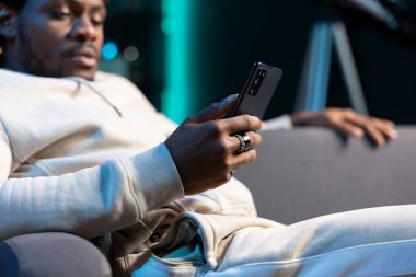 Close up shot on smartphone held by person chatting with online friends, enjoying himself at home. Focus on mobile device used by african american man in blurry background to scroll on social media clipart