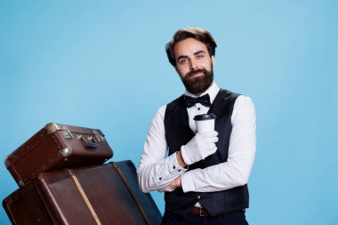 Hotel porter holding cup of coffee on camera, posing in studio dressed in formal classy attire. Young adult with bellbpy occupation drinking cold brew beverage, stands next to luggage. clipart