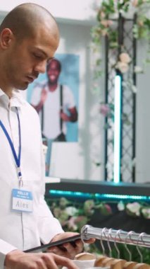 Dikey Video Perakende Asistanı, modaya uygun bir butikte raflardaki kıyafetleri inceleyerek müşteriler için ürün seçenekleri hazırlıyor. Orta Doğulu adam moda tutkusu, kaliteli tasarımcı kıyafetleri. Kamera.