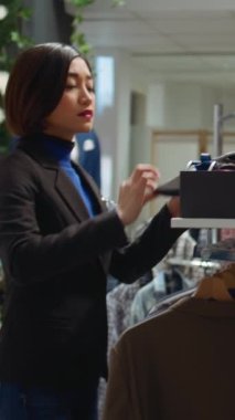 Dikey Videolu Asyalı moda aşığı çalışanı elbise mağazasındaki aksesuar reyonunda kravat ve kemer düzenliyor ve yeni ürün koleksiyonunu sergiliyor. Kıyafet zorunlulukları. Kamera B.