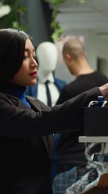 Dükkandaki yeni aksesuar koleksiyonunu sıralayan dikey video kadın stilist alışveriş merkezi çalışanı, vitrindeki kutuya kemer ve kravat yerleştirdi. Asyalı kadın butiği açmak için hazırlıyor. Kamera A