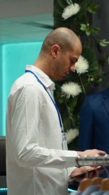 Dikey Video Dükkanı çalışanı yeni kıyafet koleksiyonu düzenliyor, ürünleri rafa koyuyor ve müşteri hizmetlerini vurguluyor. Moda bilgisi, modern perakende mağaza alanı