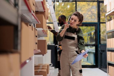 Uzaktan kumandalı bir yöneticiyle telefonda konuşan kadın denetçi depoda ürün envanterini tartışıyor. Müşteri siparişlerinde çalışan çalışanlar, karton kutuları sevkiyat için hazırlıyorlar.