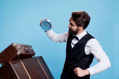 Bellhop worker looks at hour on watch, checking time while he stands against blue background. Classy doorkeeper with hotel concierge job using wall clock to see exact time. clipart