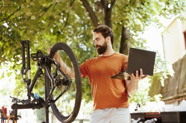Aktif erkek bisikletçi, yaz tatilinde dışarıda dizüstü bilgisayarla bisiklet sürmek için bisiklet bileşenlerinin güvenliğini arttırır. Zararı onarmak için detaylı talimatlar için elinde mini bilgisayar tutan sportif bir adam.