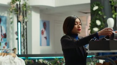 Asyalı moda aşığı işçi elbise mağazasındaki aksesuar reyonunda kravat ve kemer düzenliyor ve yeni ürün koleksiyonunu sergiliyor. Kıyafet zorunlulukları. Kamera A.
