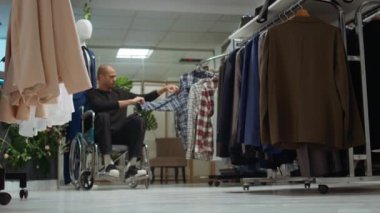 Yeni moda koleksiyonu için alışveriş yapan Ortadoğulu bir adam alışveriş merkezindeki galeriden yeni kıyafetler alıyor. Bir sürü giysi seçeneği olan tekerlekli sandalyedeki genç. Kamera B.