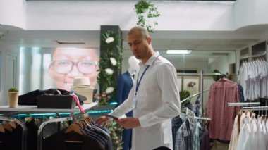Orta Doğulu perakende işçisi raflardaki yeni ürün koleksiyonunu düzenliyor, şık bir alışveriş merkezinde moda ürünleri düzenliyor. Tezgahtar detaylara dikkat ediyor. Kamera B.