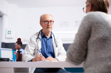 Kıdemli erkek doktor, masada oturuyor, sağlık kontrolü sırasında yaşlı kadın hastayla görüşüyor, panoya semptomlar yazıyor. Emekli doktor klinikte yaşlı bir kadınla konuşuyor..