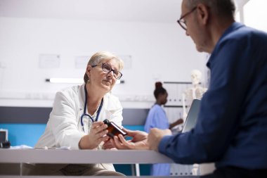 Laboratuvar önlüklü kadın doktor reçeteli ilaç tutuyor, klinikteki yaşlı erkek hastayla uyuşturucu etkilerini tartışıyor. Dost canlısı doktor, iyileşmesi için emekli hasta adama ilaç şişesi sunuyor..