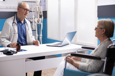 Yaşlı erkek doktor, tıbbi muayene sırasında tekerlekli sandalyedeki emekli beyaz kadına rahatlatıcı tavsiyelerde bulunuyor. Üst düzey doktor, klinikte dizüstü bilgisayarla yaşlı bayana sağlık stratejileri sunuyor..