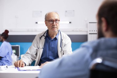 Tıbbi atamada, emekli erkek doktor genç bir adamın ortak belirtilerini dinler. Kıdemli doktor klinikteki erkek tekerlekli sandalye kullanıcısına dostça ve özenli bir danışma sunuyor..