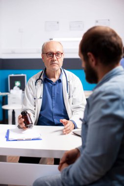 Klinikte genç beyaz adam meşgul otururken laboratuvar önlüklü yaşlı doktor ağrı kesicinin uyuşturucu etkilerini açıklıyor. Yaşlı hekim tıbbi danışmanlık sırasında erkek hastaya reçeteli ilaç veriyor..