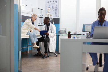 Siyahi doktor, tedavi planını özetlemek için pano kullanarak emekli beyaz kadınla sohbet ediyor. Kafkasyalı yaşlı hasta, hekim hastane odasında dikkatle dinlerken sorular soruyor..