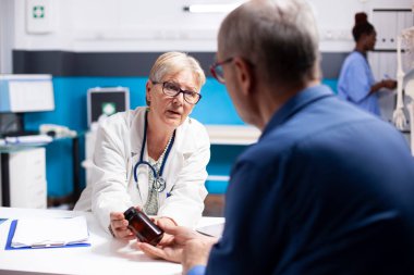 Beyaz, kıdemli bir adam masasında oturuyor. Dost canlısı bir kadın doktor, tıbbi muayene sırasında reçeteli ilaçla tedavi planını açıklıyor. Erkek emeklilik hastasına ilaç tedavisi için danışılmaktadır.