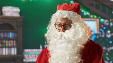 Santa Claus in his red suit browses webpages in his North Pole workshop, making sure Christmas gifts are ready for delivery. His laptop and festive surroundings highlight the holiday magic. Camera A. clipart