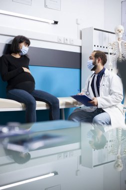 Laboratuvar önlüklü doktor, hamile bir kadının sağlık durumunu inceleyip hastanede tavsiye veriyor. Hamile bayan karnına dokunuyor ve hamilelik kontrolünde doktorla konuşuyor..