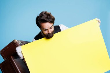 Hotel worker stands with empty poster in studio, holding blank banner with copyspace to create marketing ad campaign. Bellhop dressed in formal attire presents yellow banner on camera. clipart