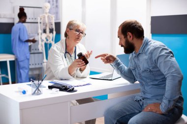 Dost canlısı yaşlı doktor, genç beyaz bir adamla sağlık sigortasını tartışırken ilaç şişesini tutuyor ve ona reçeteli ilaç tedavisinde rehberlik ediyor. Erkek hasta, kıdemli hekime ilacı hakkında danışmanlık yapıyor..